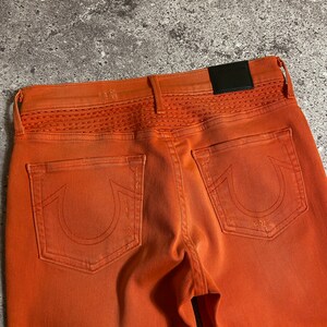 May include: A pair of orange denim jeans with a stitched detail on the back waistband and a True Religion patch on the back pocket.