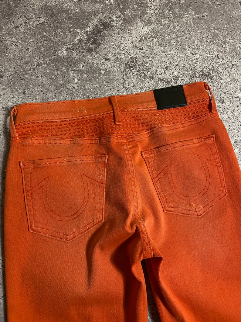 May include: A pair of orange denim jeans with a stitched detail on the back waistband and a True Religion patch on the back pocket.