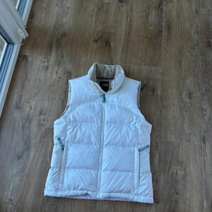 May include: A white puffer vest with a high collar and a full-length zipper. The vest has two side pockets and a small logo on the chest. The vest is made of a quilted material and is designed for warmth.