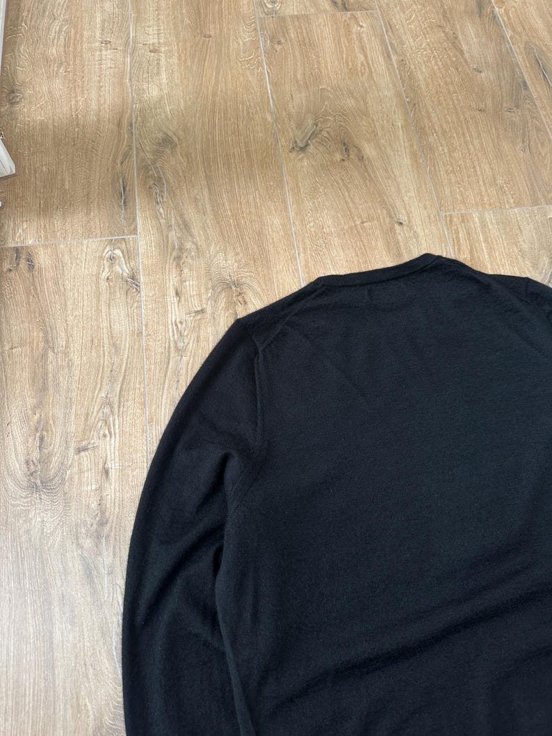Puede incluir: Un su&eacute;ter negro liso de manga larga con un cuello redondo cl&aacute;sico y una textura de punto fino. La prenda est&aacute; confeccionada con mangas montadas y un cuello acanalado, mostrando una apariencia limpia y uniforme en toda la superficie.