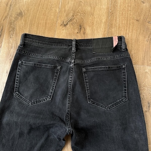 May include: A pair of black denim jeans featuring a high-rise waistband, two back pockets, and a black leather-like patch on the rear right side with a small pink accent. The jeans have visible stitching details and a classic five-pocket construction.