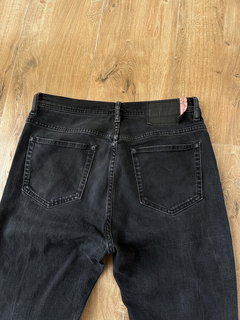May include: A pair of black denim jeans featuring a high-rise waistband, two back pockets, and a black leather-like patch on the rear right side with a small pink accent. The jeans have visible stitching details and a classic five-pocket construction.