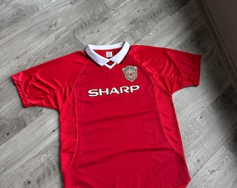 May include: Red short-sleeved jersey with a white collar and the word "SHARP" printed across the chest. A team crest is on the upper left chest. The jersey is lying on a wooden floor.