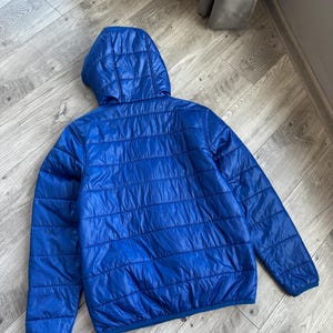 May include: A bright blue, quilted jacket with a hood. The jacket has a horizontal, stitched pattern and a smooth, shiny finish. The back view shows the full jacket, laid flat.