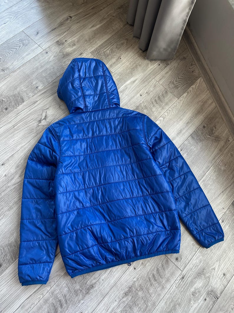 May include: A bright blue, quilted jacket with a hood. The jacket has a horizontal, stitched pattern and a smooth, shiny finish. The back view shows the full jacket, laid flat.