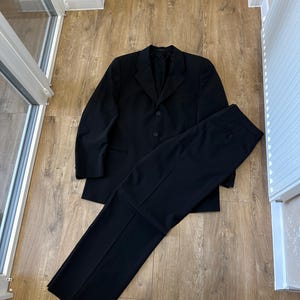以下が含まれることがあります： ノッチドラペルと2つのフロントボタンを備えたシングルブレストのブレザーと、お揃いのストレートレッグパンツで構成される無地の黒いツーピーススーツ。長袖で、すっきりとした構造的なデザインが特徴です。
