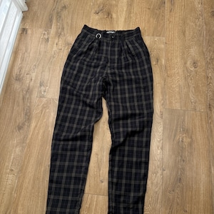 May include: High-waisted trousers in a black and dark brown plaid pattern. These pants feature a front button closure, a small silver-toned metal ring attached to the waistband, and subtle pleats below the waistline, tapering toward the hem.