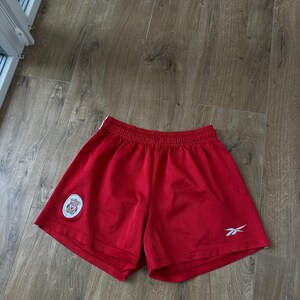 May include: Red athletic shorts with a white logo on the left leg and a small white Reebok logo on the right leg. The shorts have a white stripe down the side and an elastic waistband. The shorts are made of a breathable material.