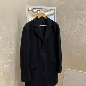 May include: A black overcoat hanging on a wooden hanger in front of a mirror. The coat has a classic design with a notched lapel, front pockets, and button closures. The coat is a solid black color.