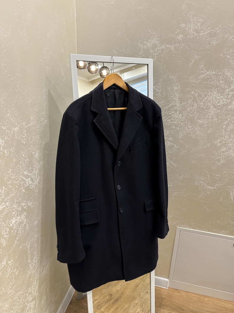 May include: A black overcoat hanging on a wooden hanger in front of a mirror. The coat has a classic design with a notched lapel, front pockets, and button closures. The coat is a solid black color.