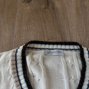 Puede incluir: C&aacute;rdigan de punto trenzado color crema con cuello en V y ribete marr&oacute;n. El cuello y los bordes presentan rayas en marr&oacute;n, beige y azul. El nombre de la marca "Falconeri" es visible en una peque&ntilde;a etiqueta.