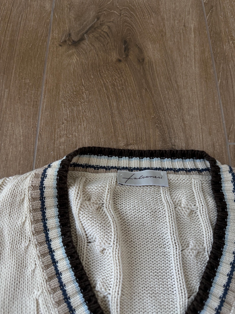 Puede incluir: C&aacute;rdigan de punto trenzado color crema con cuello en V y ribete marr&oacute;n. El cuello y los bordes presentan rayas en marr&oacute;n, beige y azul. El nombre de la marca "Falconeri" es visible en una peque&ntilde;a etiqueta.