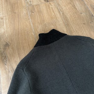 Op de afbeelding: Een donker antracietgrijs gebreid jasje met een effen zwarte geribbelde opstaande kraag. Het kledingstuk is gemaakt van een getextureerde gebreide stof met zichtbare naden die verticaal over de rug en langs het schoudergedeelte lopen.