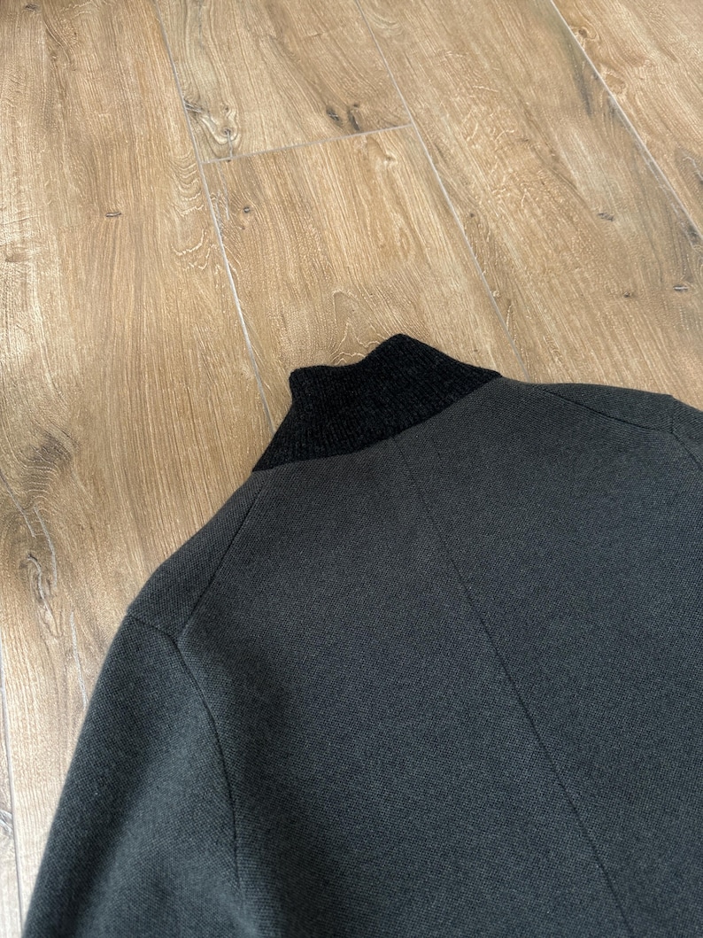 Op de afbeelding: Een donker antracietgrijs gebreid jasje met een effen zwarte geribbelde opstaande kraag. Het kledingstuk is gemaakt van een getextureerde gebreide stof met zichtbare naden die verticaal over de rug en langs het schoudergedeelte lopen.