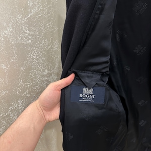 May include: A dark navy blue coat with a black interior lining. The interior lining features a repeating pattern and a label that reads "Boggi Milano". The coat's interior pocket is visible.