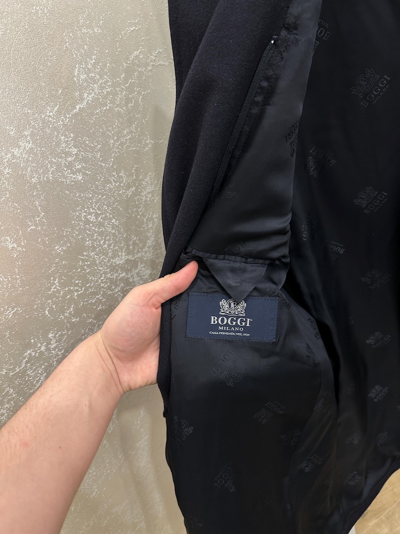 May include: A dark navy blue coat with a black interior lining. The interior lining features a repeating pattern and a label that reads "Boggi Milano". The coat's interior pocket is visible.