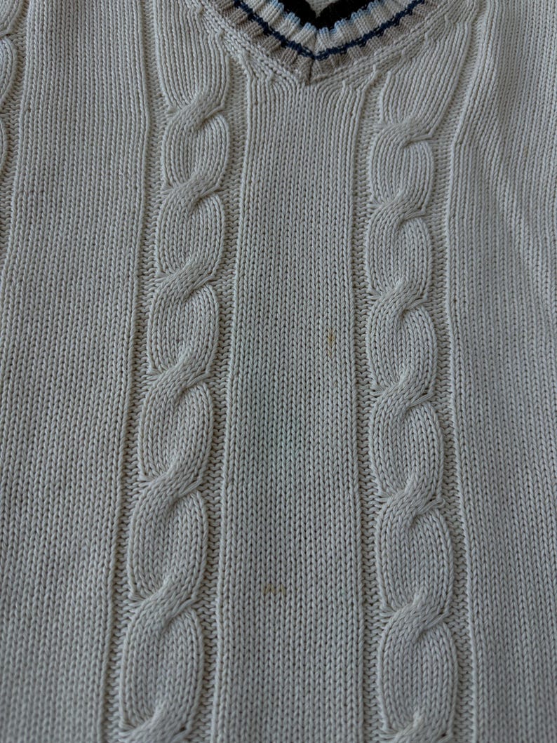 Puede incluir: Su&eacute;ter de punto trenzado color crema con cuello en V. El su&eacute;ter presenta un dise&ntilde;o de punto trenzado texturizado en la parte delantera y un ribete a rayas en el cuello. La prenda de punto es de color neutro, adecuada para varios conjuntos.