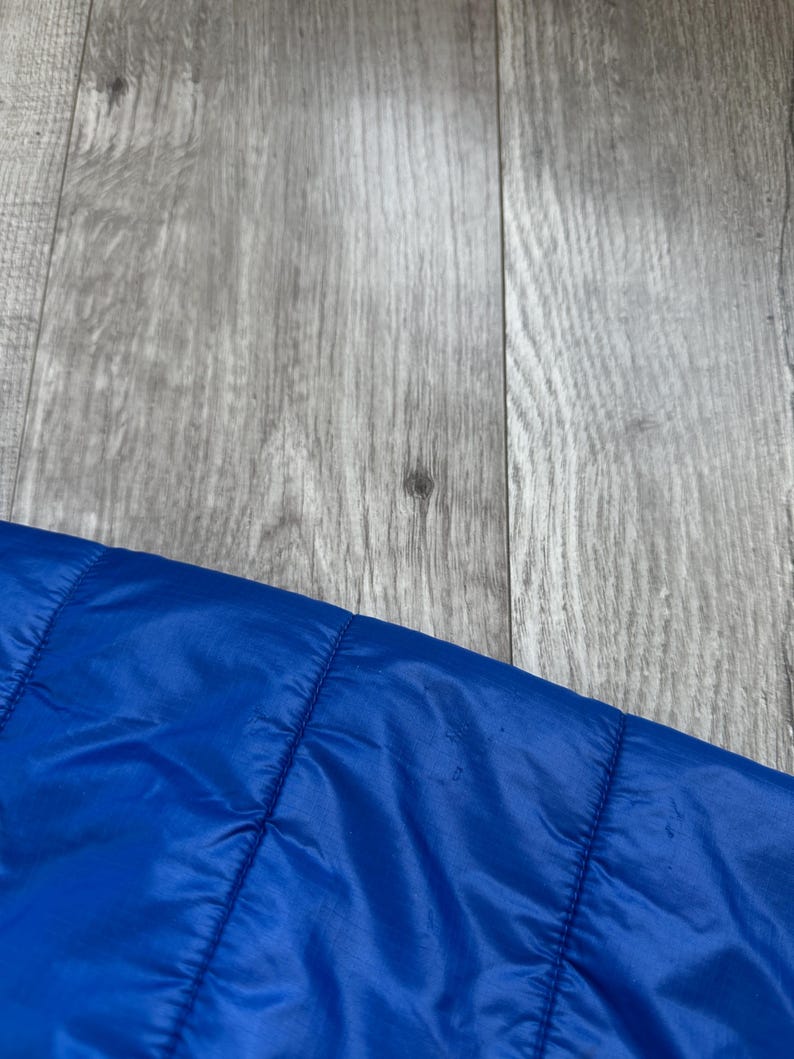 May include: Close-up of a section of a bright blue quilted jacket. The jacket has a shiny, smooth texture and is stitched with horizontal lines. The background is a gray wooden surface.