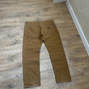 Puede incluir: Pantalones de trabajo marrones con diseño de pernera recta. Los pantalones tienen dos bolsillos traseros y un pequeño parche cuadrado en el bolsillo trasero derecho. Los pantalones están hechos de un material duradero.