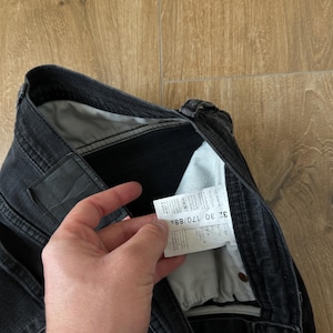 May include: Black denim jeans featuring a white interior care label with the text 32 30 170/88A printed clearly. The jeans include a dark waistband, metal rivets, and a light grey pocket lining visible inside the garment construction.
