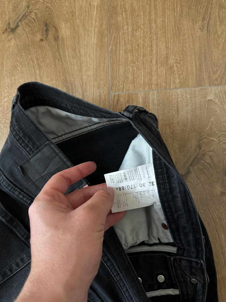 May include: Black denim jeans featuring a white interior care label with the text 32 30 170/88A printed clearly. The jeans include a dark waistband, metal rivets, and a light grey pocket lining visible inside the garment construction.