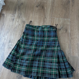 May include: A green, blue, and black plaid kilt. The kilt is made of a woven fabric with a traditional tartan pattern. The kilt has pleats and a leather belt. The kilt is approximately 24 inches long.