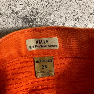 May include: A close-up of the inside of a pair of orange jeans with a brown leather tag that reads "HALLE Mid Rise Super Skinny" and a brown leather tag that reads "TRUE RELIGION 26 MADE IN USA OF IMPORTED FABRIC FABRIQUE AUX ETATS-UNIS".