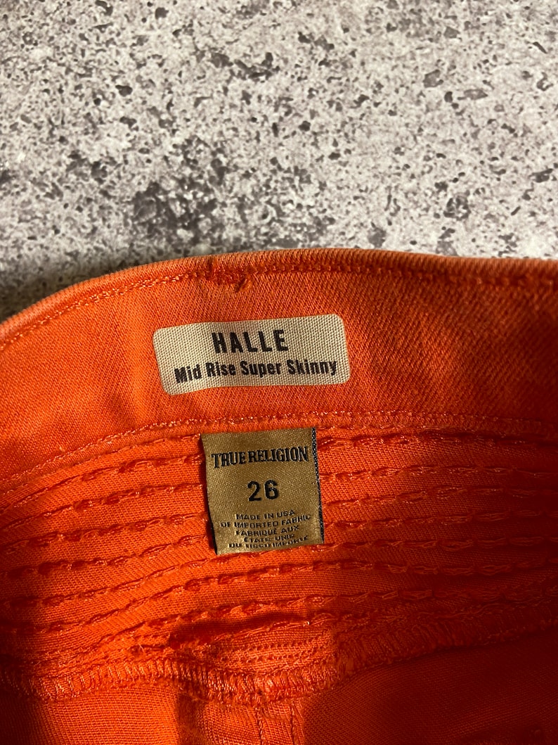 May include: A close-up of the inside of a pair of orange jeans with a brown leather tag that reads "HALLE Mid Rise Super Skinny" and a brown leather tag that reads "TRUE RELIGION 26 MADE IN USA OF IMPORTED FABRIC FABRIQUE AUX ETATS-UNIS".