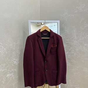 Puede incluir: Un blazer de color burdeos intenso con solapa de muesca, dos botones oscuros y dos bolsillos delanteros de parche. Un pañuelo de bolsillo rojo oscuro con estampado de lunares blancos se encuentra en el bolsillo del pecho. El forro es gris oscuro.