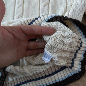 Puede incluir: Su&eacute;ter de punto trenzado color crema con un dobladillo acanalado con rayas marrones, azul claro y crema. El su&eacute;ter tiene una etiqueta que dice "BABY COTTON" y el n&uacute;mero "54".