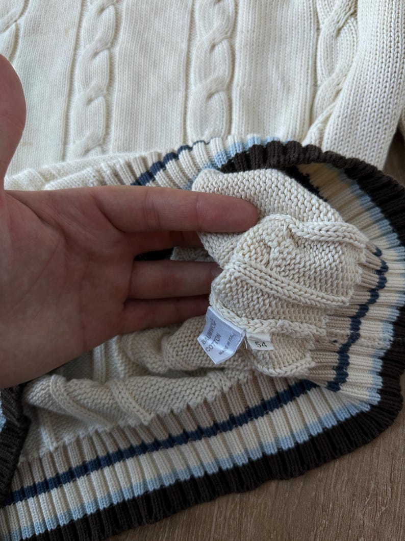 Puede incluir: Su&eacute;ter de punto trenzado color crema con un dobladillo acanalado con rayas marrones, azul claro y crema. El su&eacute;ter tiene una etiqueta que dice "BABY COTTON" y el n&uacute;mero "54".