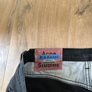 May include: A pair of black denim jeans featuring a rectangular pink fabric label sewn onto the interior waistband. The label displays the text Acne Bl&aring; Konst, Floragatan 13, Stockholm, and Studios in black and blue lettering against a pink background.