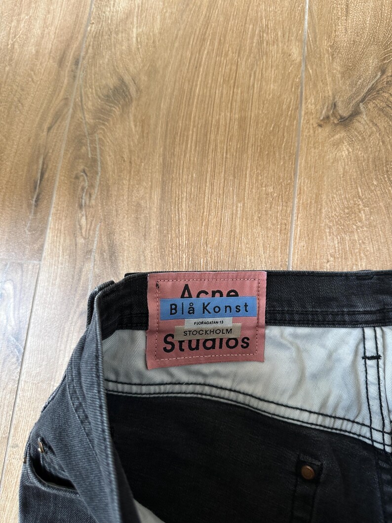 May include: A pair of black denim jeans featuring a rectangular pink fabric label sewn onto the interior waistband. The label displays the text Acne Bl&aring; Konst, Floragatan 13, Stockholm, and Studios in black and blue lettering against a pink background.