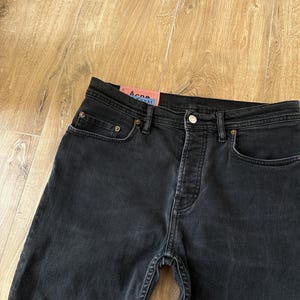 May include: Black denim jeans featuring a button fly closure, five-pocket styling with copper-toned rivets, and a pink rectangular brand label on the interior waistband with the text Acne Studios Konst printed in black and blue lettering.