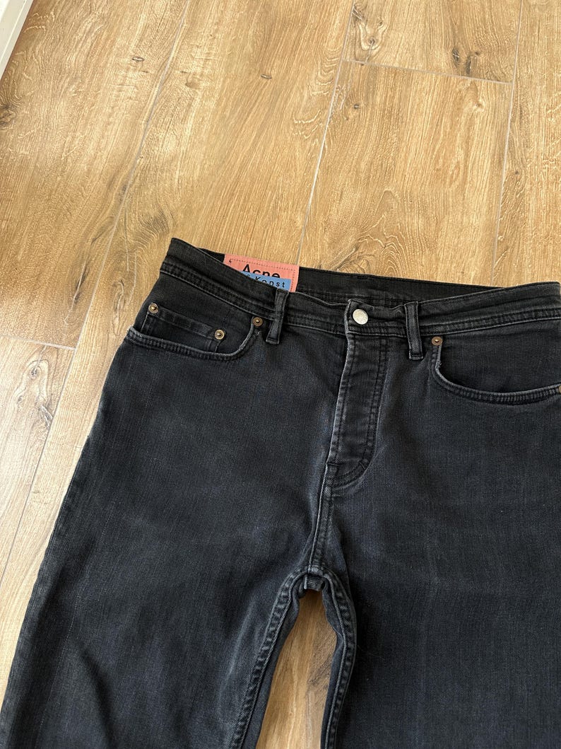 May include: Black denim jeans featuring a button fly closure, five-pocket styling with copper-toned rivets, and a pink rectangular brand label on the interior waistband with the text Acne Studios Konst printed in black and blue lettering.