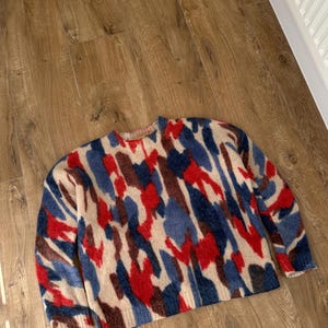 May include: A long-sleeved crew neck sweater featuring a fuzzy, brushed texture. The knit garment displays an abstract, multi-colored pattern consisting of bold splashes of red, dark brown, and muted blue set against a light beige background.