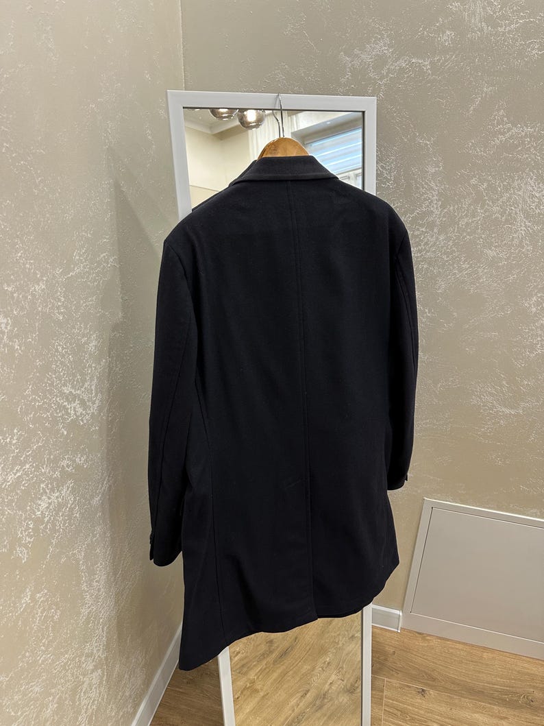 May include: A dark navy blue coat hanging on a wooden hanger inside a white framed mirror. The coat is a classic style with a tailored fit and a single-breasted design. The coat is made of a smooth, solid fabric.