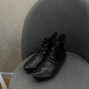 May include: A pair of black leather ankle boots featuring a pointed toe, a lace-up front closure with thin black laces, and a structured collar that extends slightly upward at the back. The boots have a smooth, matte finish and a low-profile sole.