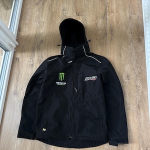 May include: A black softshell jacket featuring a hood with drawstrings, a full-length front zipper, and long sleeves with adjustable cuffs. The chest displays a green Monster Energy logo on the right and a white Bud Racing logo on the left.