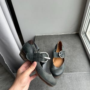 May include: A pair of dark teal Mary Jane style shoes with a low heel. The shoes feature a strap with a silver buckle and a leather sole. The shoes are displayed on a gray surface.