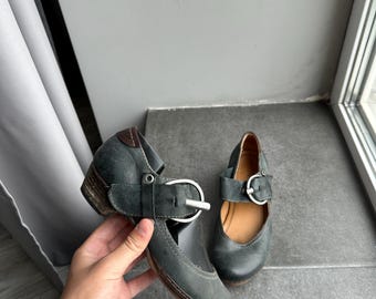 May include: A pair of dark teal Mary Jane style shoes with a low heel. The shoes feature a strap with a silver buckle and a leather sole. The shoes are displayed on a gray surface.