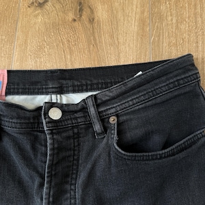 May include: A pair of black denim jeans featuring a button fly closure with a silver-toned metal button, belt loops, and a small coin pocket. A pink rectangular fabric tag is attached to the waistband on the left side of the garment.