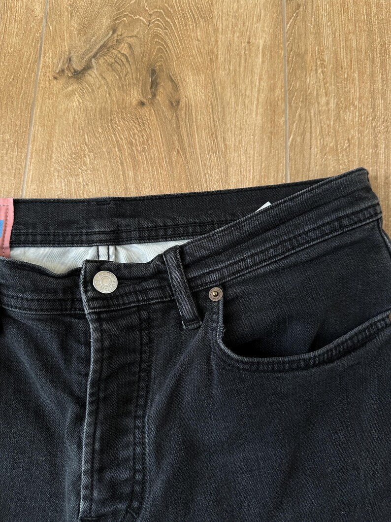 May include: A pair of black denim jeans featuring a button fly closure with a silver-toned metal button, belt loops, and a small coin pocket. A pink rectangular fabric tag is attached to the waistband on the left side of the garment.