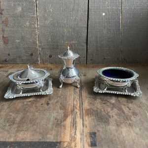 May include: Three silver salt and pepper shakers with ornate designs. The shakers are on a wooden surface.