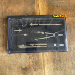 May include: A set of silver drafting tools, including compasses and dividers, neatly arranged in a black plastic case. The case has a clear lid and the brand name "Skala P III" is visible. The tools are on a wooden surface.