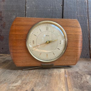 May include: A vintage Smiths clock with a light wood frame and a cream-colored face. The clock has gold-tone hands and a gold-tone bezel. The clock face displays the word "Smiths". The clock is supported by a gold-tone metal base.