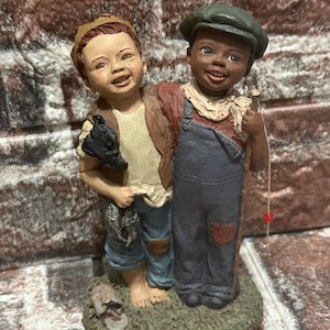 Può includere: Una statuina raffigurante due ragazzi, uno con la pelle chiara e l'altro con la pelle scura, in piedi insieme. Il ragazzo con la pelle chiara tiene una scarpa e l'altro una canna da pesca. Sono in piedi su un ciuffo d'erba.