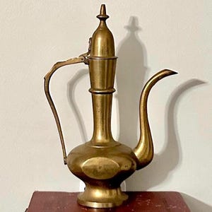 May include: A vintage brass teapot with a long spout and handle, resting on two antique books. The teapot has a decorative design and a pointed top. The books are red and black, adding a touch of classic style to the scene.