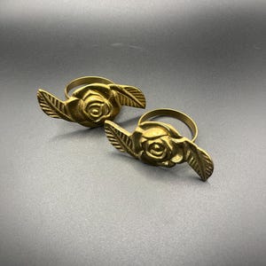 May include: Two brass-colored napkin rings. Each ring features a rose in the center with two leaf accents. The rings are made of metal and are set against a dark gray background. The rings are designed to hold cloth napkins.