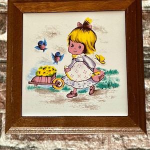 May include: A framed tile with a painted image of a girl with yellow hair and a pink dress pushing a wheelbarrow full of yellow flowers. Two blue birds are flying in the background. The tile is surrounded by a brown wooden frame.
